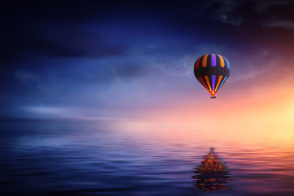 Lautlos gleitet eine prächtige, farbenfrohe Montgolfiere über ruhige Gewässer, von sicherer Hand gesteuert. Die Silhouette des Heißluftballons reflektiert sich kontrastreich an der Wasseroberfläche; blaue sowie rot-gelbe Farbtöne dominieren.