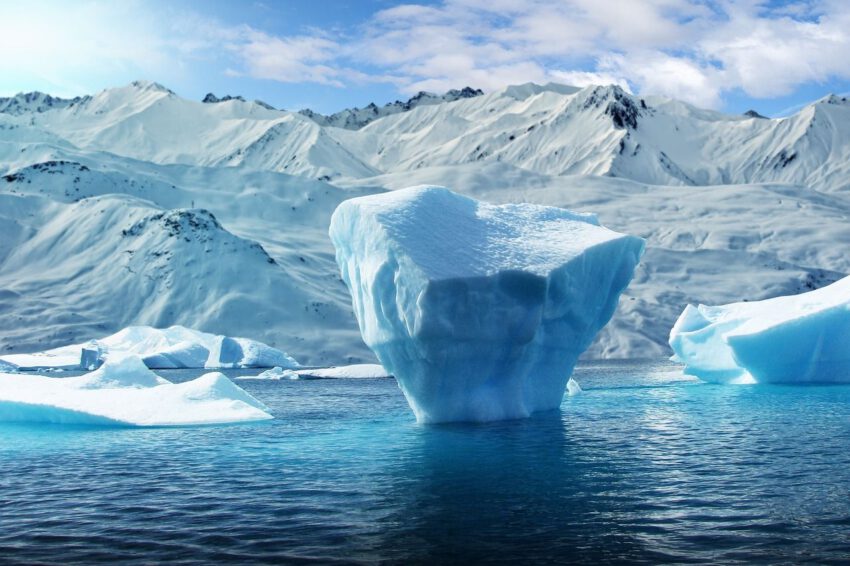 Ein Eisberg im Wasser, vor der Kulisse einer schneebedeckten Arktis-Landschaft. Die Farben blau und weiß dominieren.